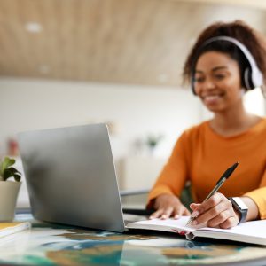 Black woman sitting at desk, using computer writing in notebook