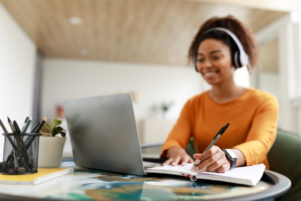 Black woman sitting at desk, using computer writing in notebook