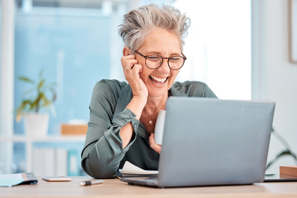 Laptop, coffee and senior business woman laughing at an email in her office at work for fun or humo